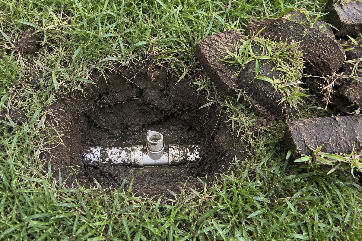 Hand-dug hole in turf exposing a lateral pipe fitting with the snapped stub of a threaded riser still in it
