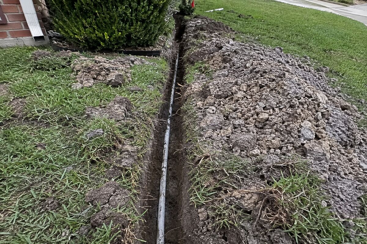 Narrow open trench in a lawn with a section of white PVC lateral pipe exposed and damp soil piled alongside