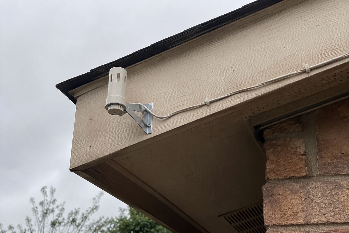 Small cylindrical rain sensor bracketed to a roof eave with its thin wire running along the fascia
