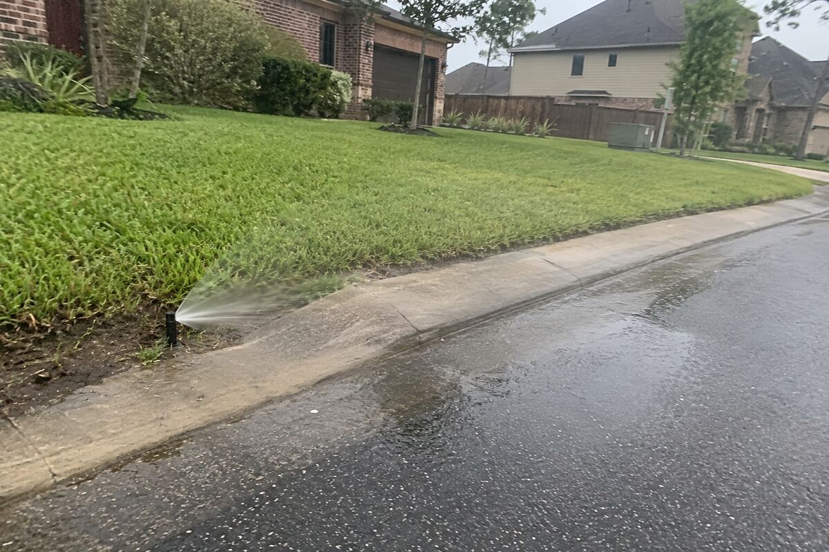 Pop-up spray head at the edge of a lawn throwing water across a concrete curb onto the roadway